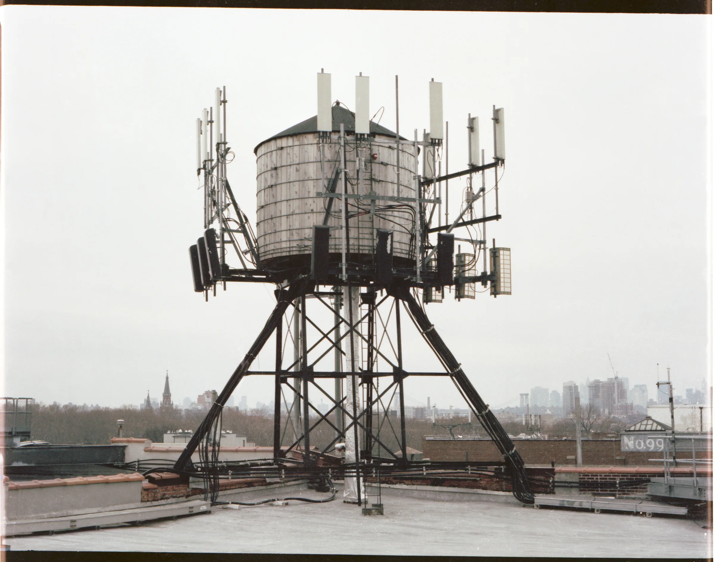 A water tower on a steel frame bristling with cell antennas, on a rooftop above the city under a white sky